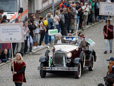 Ein Oldtimer fährt auf einem Umzug durch eine Stadt, begleitet von vielen Zusehenden.