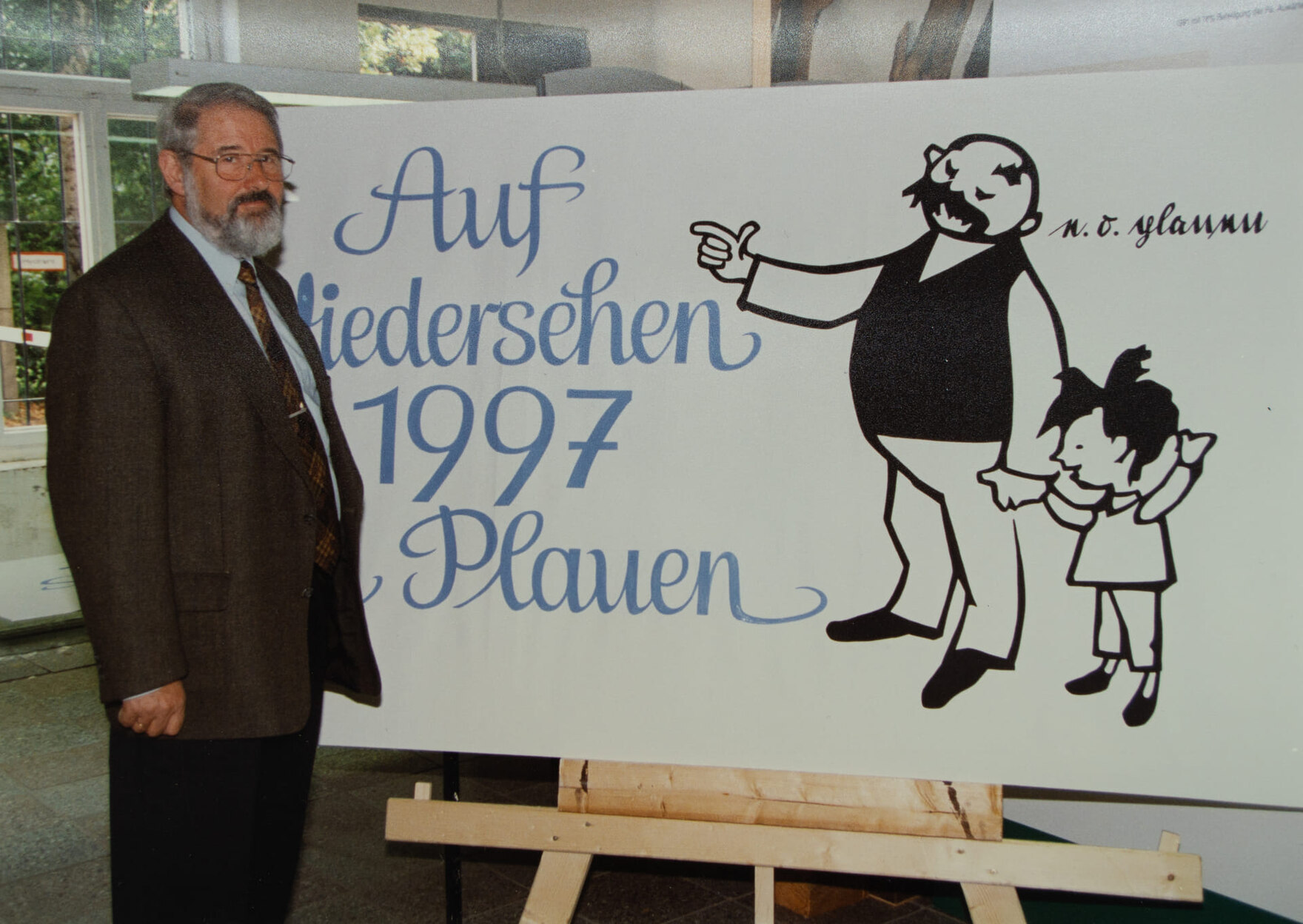 Ein Mann steht neben einer Tafel mit der Aufschrift „Auf Wiedersehen 1997 Plauen“ sowie einer gezeichneten Figurengruppe, bestehend aus einem Mann und einem Kind