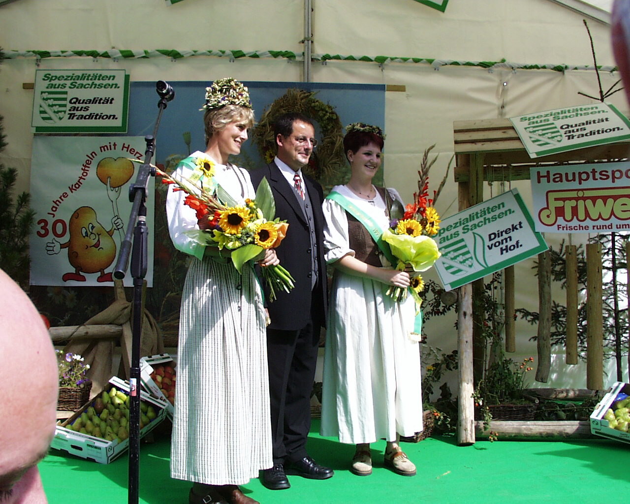 Zwei Frauen in Tracht mit Schärpen und Blumen posieren mit einem Mann auf der Bühne
