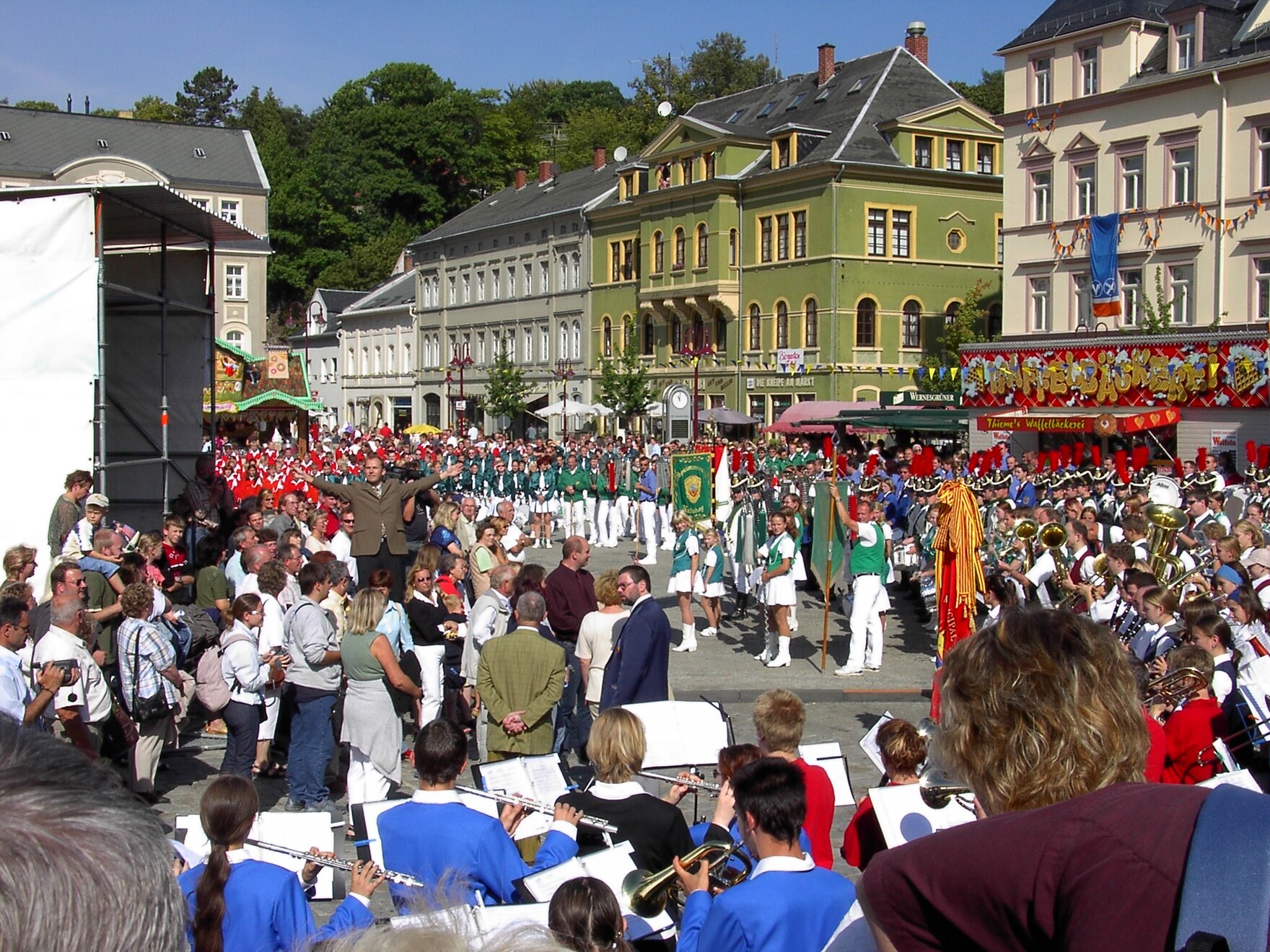 Großes Publikum auf einem belebten Stadtplatz beobachtet einen Festumzug mit Spielmannszügen