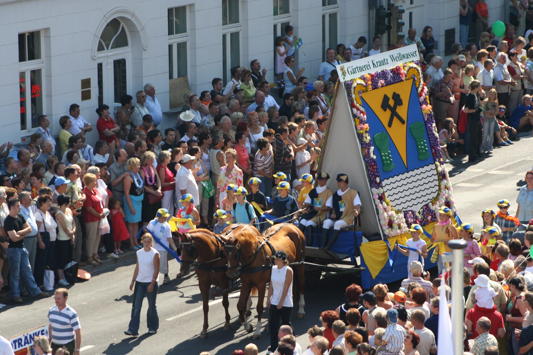 Ein von zwei braunen Pferden gezogener, mit Blumen geschmückter Festwagen mit großem Wappen fährt an einer dicht gedrängten Menschenmenge vorbei.