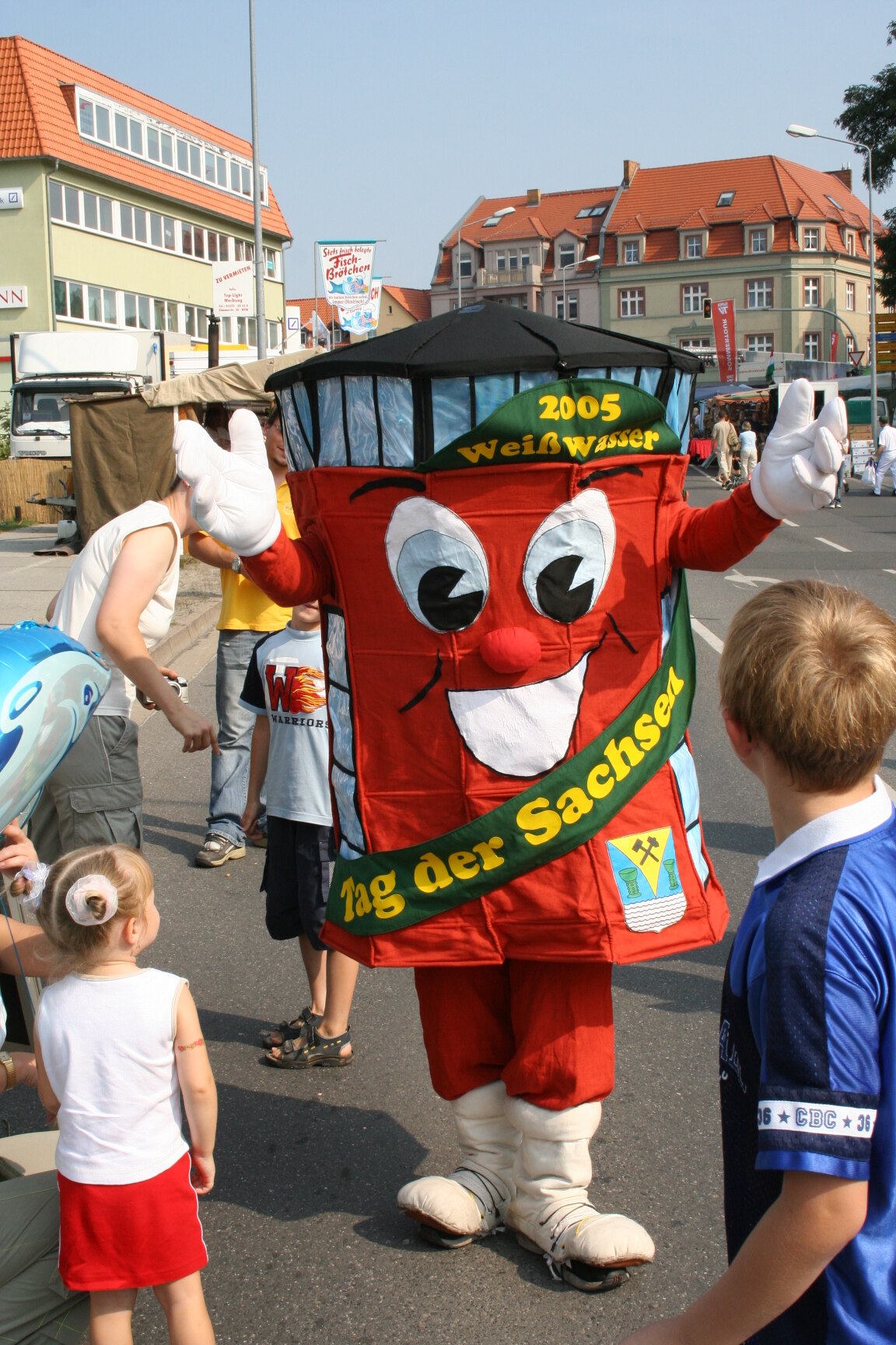 Eine Person in einem roten, lächelnden Maskottchenkostüm mit der Aufschrift „2005 Weißwasser“ und „Tag der Sachsen“ steht auf einer Straße, umgeben von Kindern