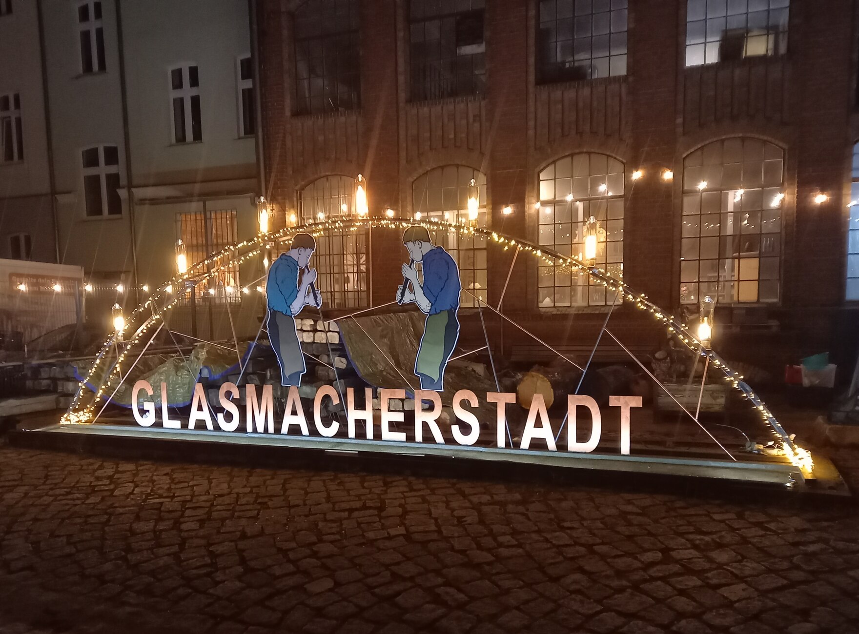 Eine beleuchtete Installation mit der Aufschrift „Glasmacherstadt“ und zwei stilisierten Glasbläserfiguren vor einer Backsteinfassade bei Nacht.