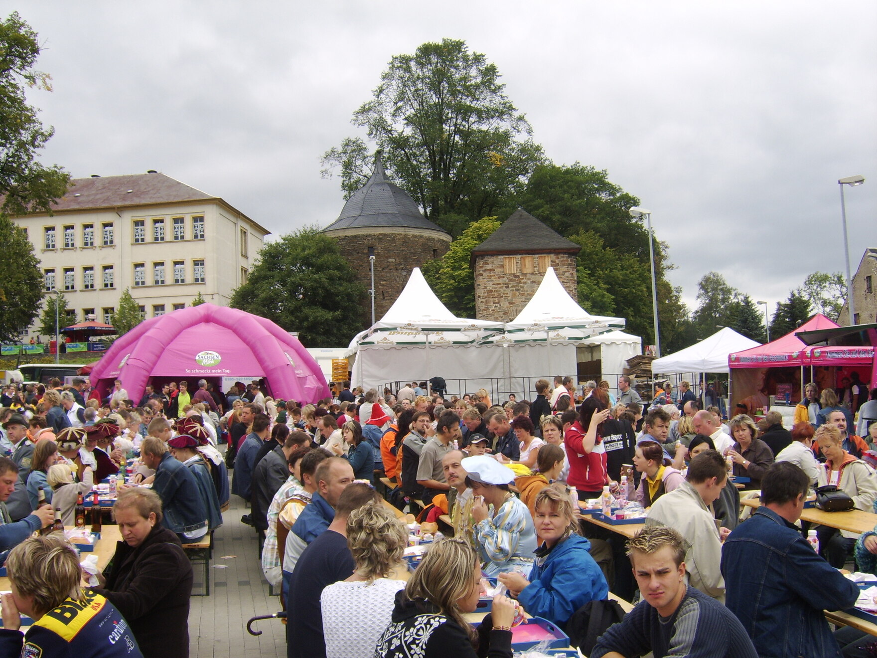 Viele Menschen sitzen an Biertischgarnituren im Freien, umgeben von bunten Pavillons