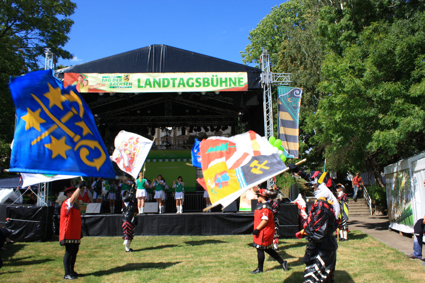 Bühne mit der Aufschrift „Landtagsbühne“ und davor kostümierte Personen, die bunte Fahnen schwenken.
