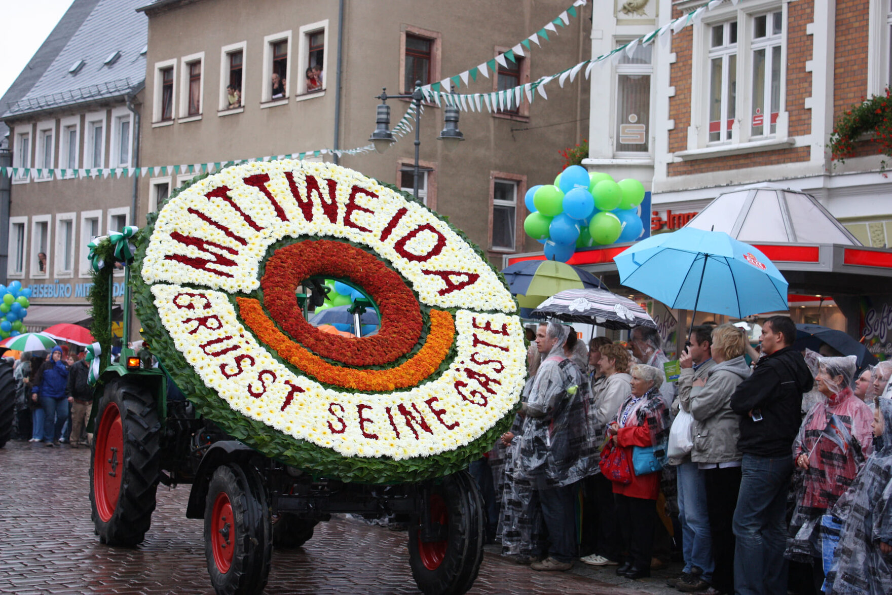 Ein mit Blumen geschmückter Wagen mit der Aufschrift „Mittweida grüßt seine Gäste“ fährt an einer Menschenmenge mit Regenschirmen vorbei