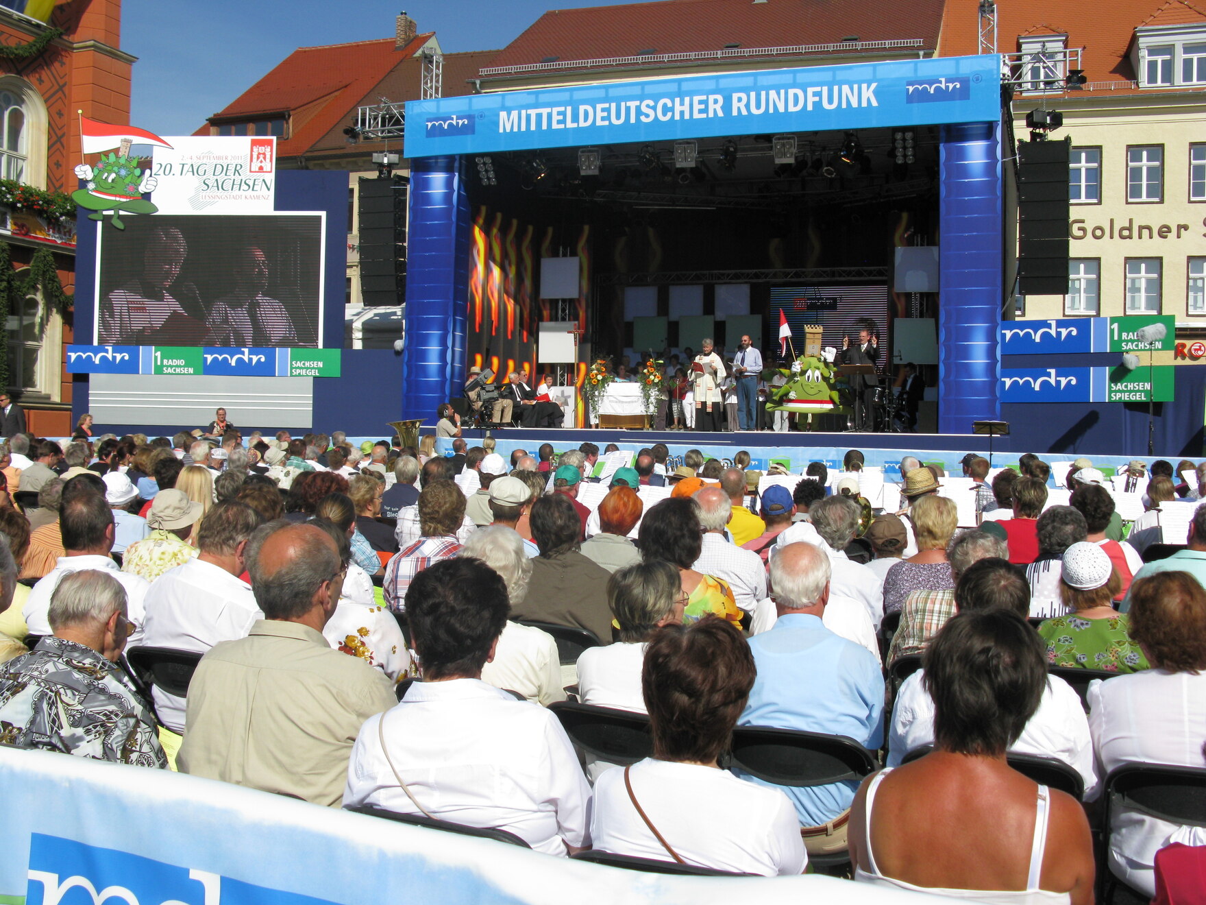 Eine Menschenmenge sitzt vor einer Bühne mit der Aufschrift „Mitteldeutscher Rundfunk“, auf der mehrere Personen und ein grünes Maskottchen stehen