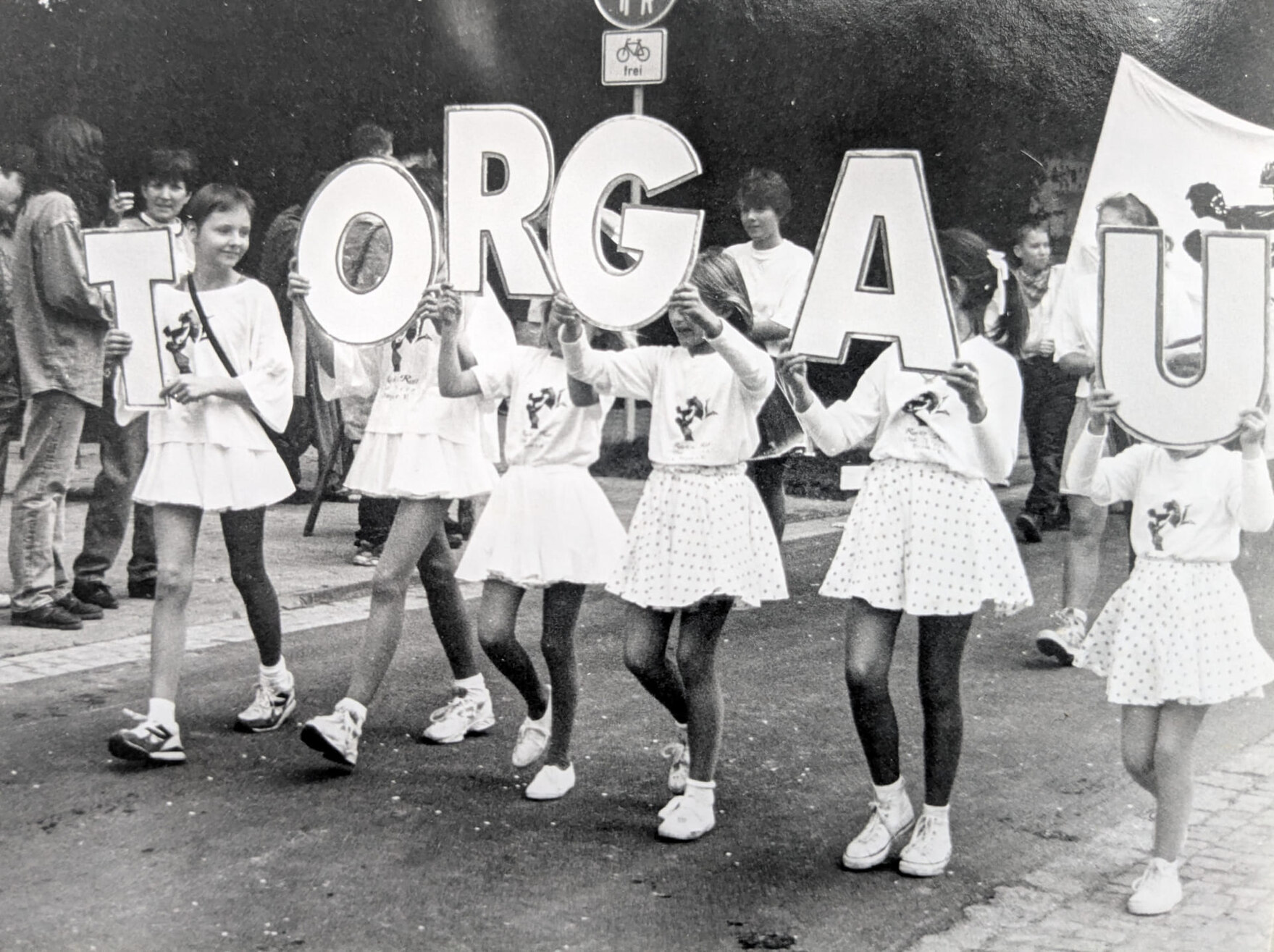 Mehrere Mädchen in einheitlicher Kleidung halten große Buchstaben hoch, die zusammen das Wort „Torgau“ ergeben, während sie in einer Parade laufen