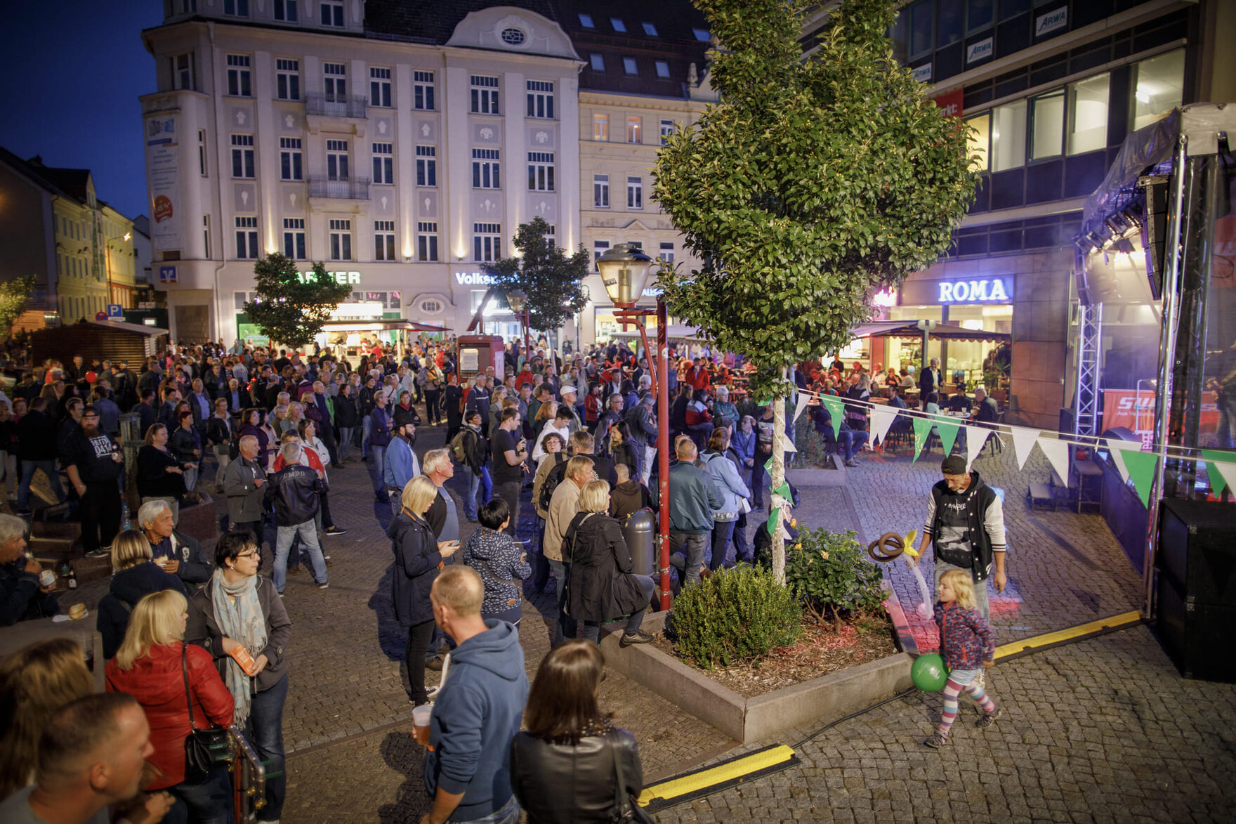 Abendliche Straßenveranstaltung mit vielen Menschen auf einem belebten Platz vor beleuchteten Gebäuden