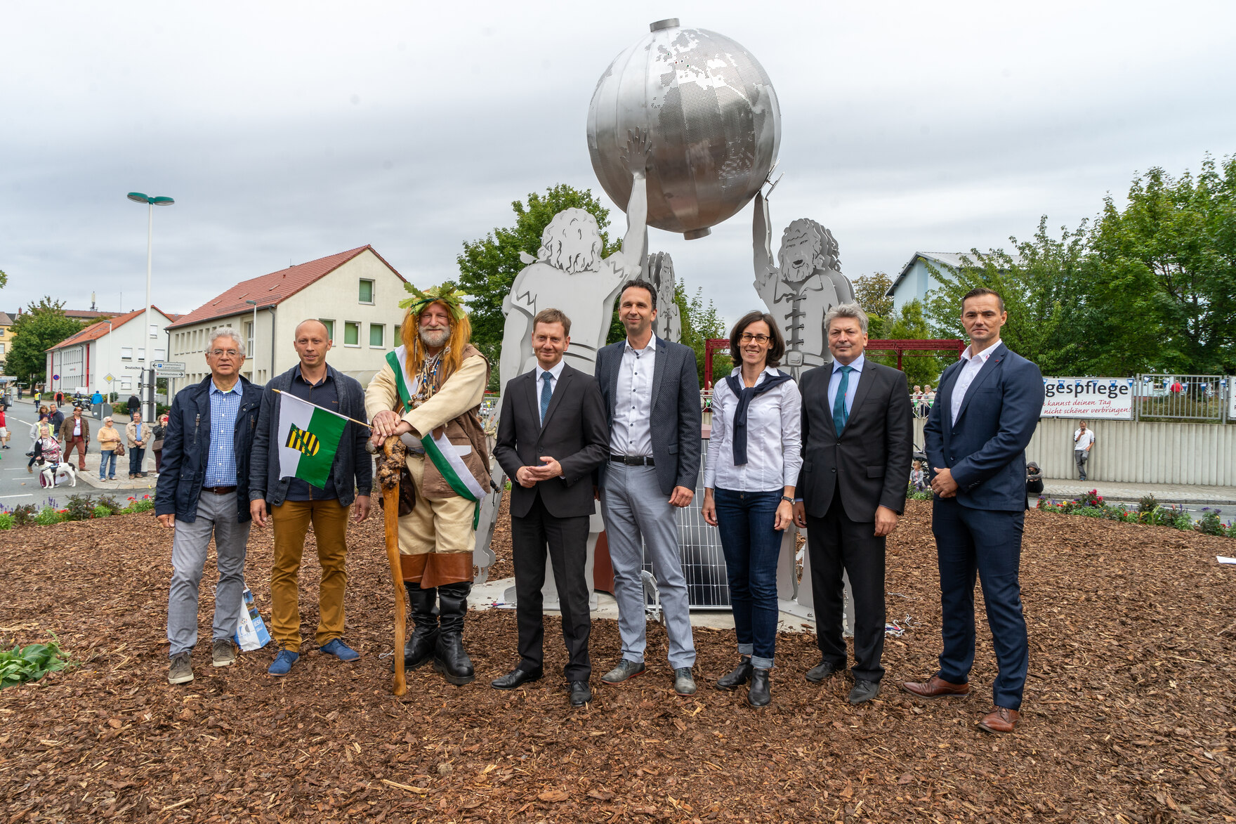 Acht Personen stehen vor einer silberfarbenen Skulptur mit stilisierten Figuren, die eine große Kugel halten