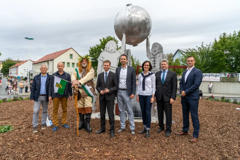 Acht Personen stehen vor einer silberfarbenen Skulptur mit stilisierten Figuren, die eine große Kugel halten